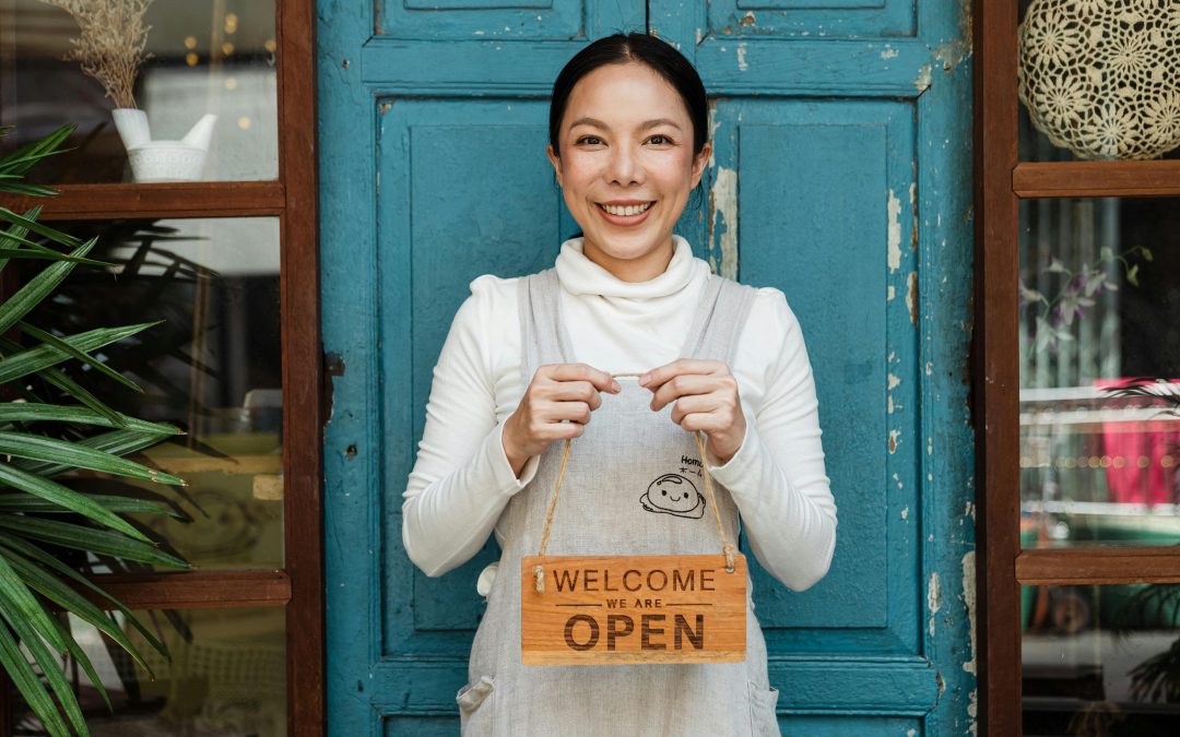 Smiling woman holding a "Welcome We Are Open" sign in front of a blue door, representing small business readiness and customer engagement.